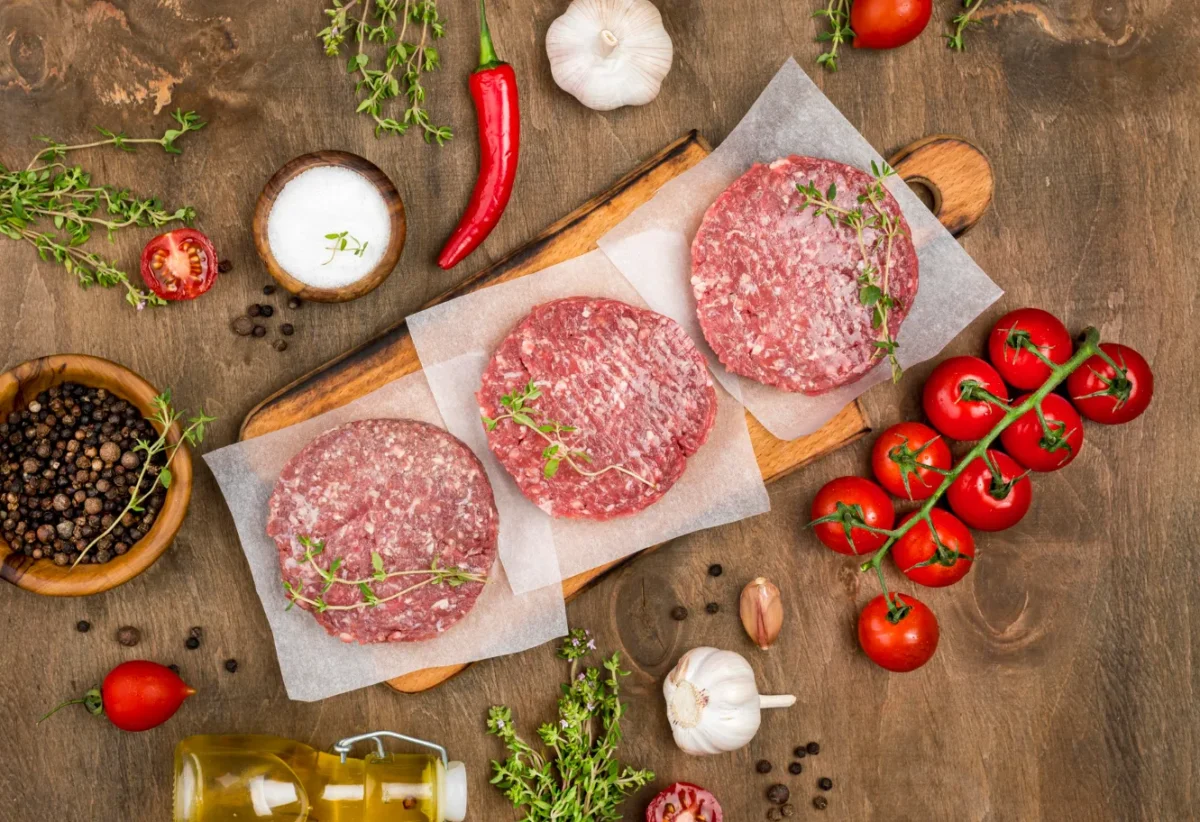 Tres hamburguesas de carne cruda sobre papel encerado, rodeadas de ingredientes frescos como tomates cherry, ajo, guindilla, sal, pimienta negra y hierbas aromáticas, sobre una tabla de madera. Ideal para recetas de cocina y preparación de comidas.