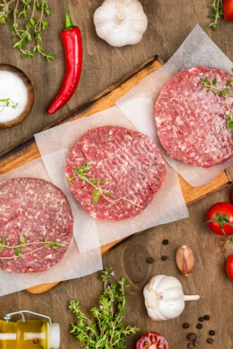 Tres hamburguesas de carne cruda sobre papel encerado, rodeadas de ingredientes frescos como tomates cherry, ajo, guindilla, sal, pimienta negra y hierbas aromáticas, sobre una tabla de madera. Ideal para recetas de cocina y preparación de comidas.
