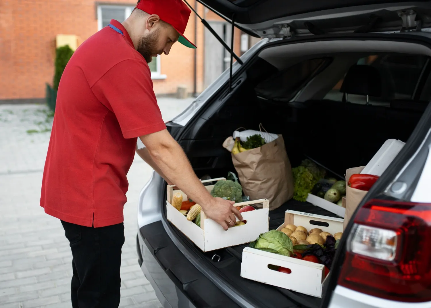Hombre cargando cajas de verduras y frutas frescas en el maletero de un coche, mostrando la importancia de la alimentación saludable y la compra de productos locales.