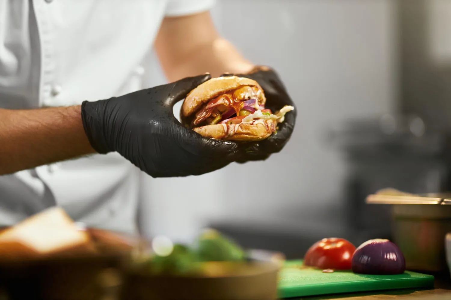 Mano con guante negro sosteniendo una hamburguesa con vegetales y salsa, en un entorno de cocina profesional.